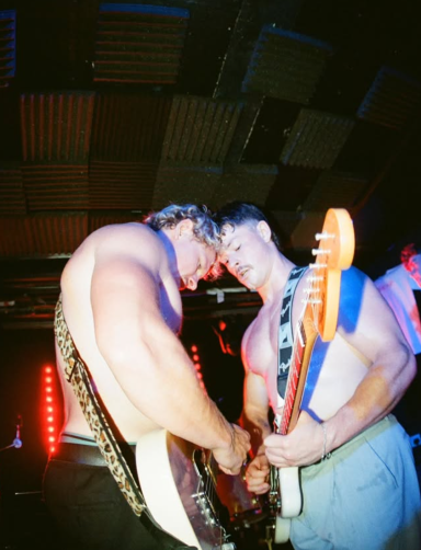FAT SALAD Episode Three Two muscular men playing electric guitars on stage under bright lights.