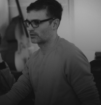 Brett Shaw Episode Five A man in glasses, focused on a task, with a blurred background of musical instruments.